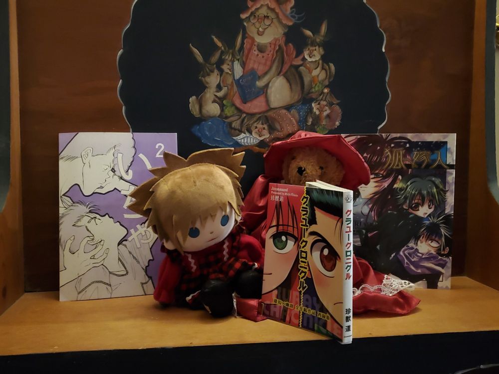 three boys' love doujinshis placed on a bench with a sora from kingdom hearts plush and a bear plush wearing a red dress